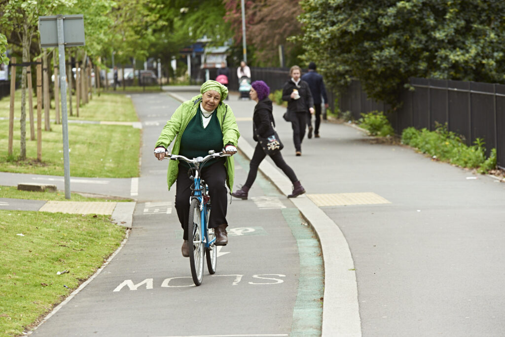 An elderly woman wearing a green coat and headscarf is riding a bicycle on a designated cycling path in an urban park. The path is marked with cycling symbols, and the surroundings include green lawns, trees, and a few pedestrians in the background.