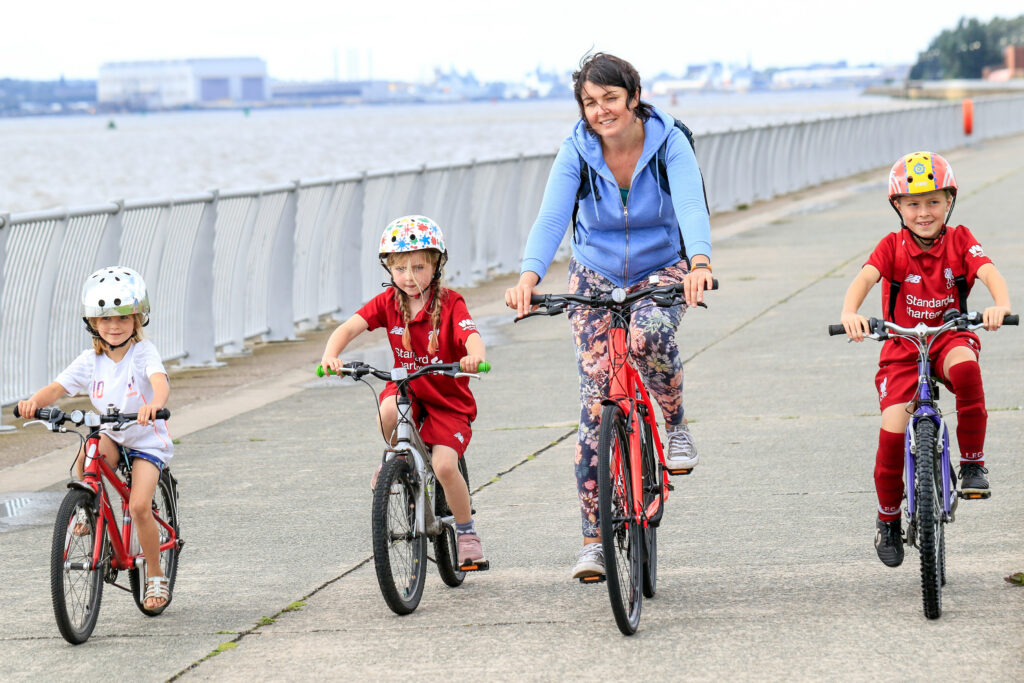 A woman rides a red bike along a waterfront path, accompanied by three children on their own bicycles. The children, wearing helmets, are dressed in football kits, and they all ride in a line, enjoying the outdoor activity.