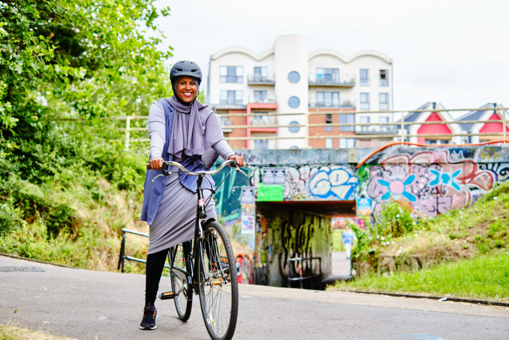 A smiling woman rides a bicycle on a sunny day along a path lined with greenery and trees. She is dressed casually, wearing a black top and patterned pants, with the clear blue sky above her.