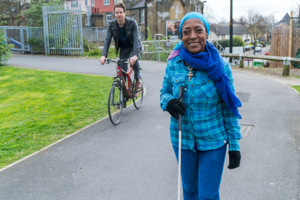 A woman wearing a blue plaid shirt, gloves, and a headscarf is smiling as she walks with a white cane. A man is riding a red bicycle behind her on a paved path in a park, with green grass and trees surrounding them.