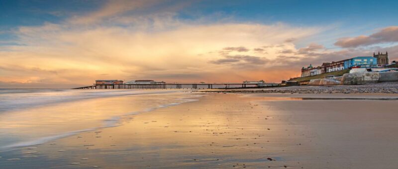 Cromer_Pier at sunset extending into the sea, reflecting warm tones in the wet sand, and a row of buildings along the shore