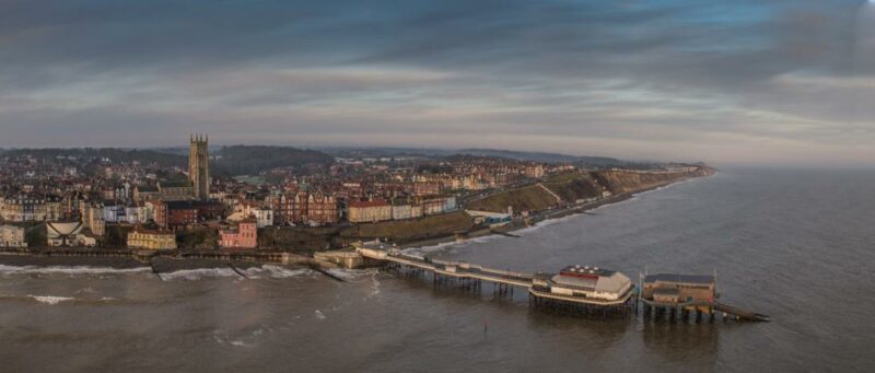 A panoramic view of Cromer_Pier with a pier extending into the sea, historic buildings, and rolling hills under a cloudy sky