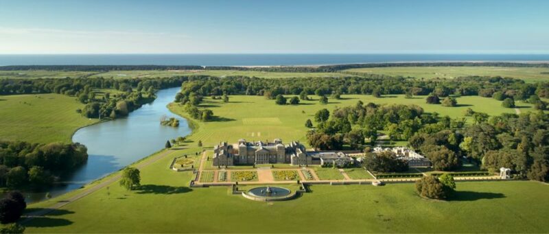 Aerial view of Holkham estate with expansive green lawns, a river, and formal gardens, stretching towards the sea under a clear blue sky.